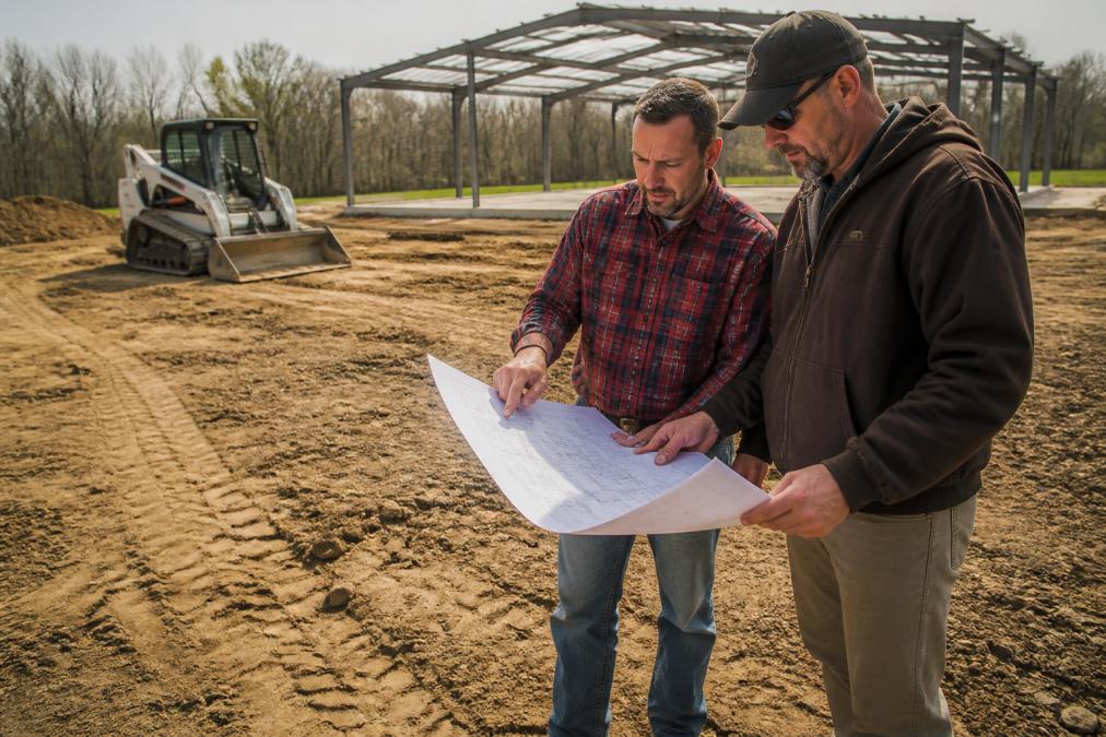 A rural property owner in a plaid shirt and work boots reviewing a printed site plan with a contractor next to a freshly graded dirt pad, skid steer parked behind them, metal building frame visible in the distance