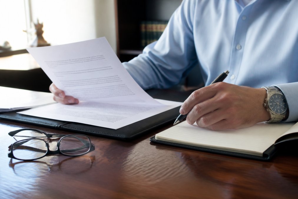 Close-up of a person reviewing a written fee agreement document at a desk with a pen, notepad, and reading glasses visible, natural office lighting
