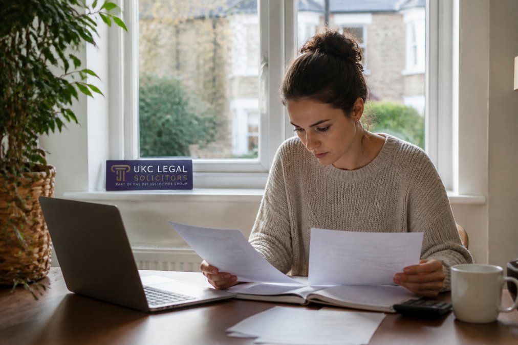 First-time homebuyer sitting at a desk with documents and a laptop, reviewing conveyancing quotes with a focused expression, natural office lighting streaming through a window