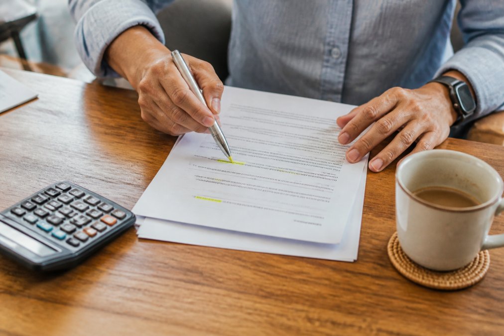 Close-up of a committee member's hands highlighting clauses in a printed strata management contract with a pen, a calculator and a coffee cup on a wooden table