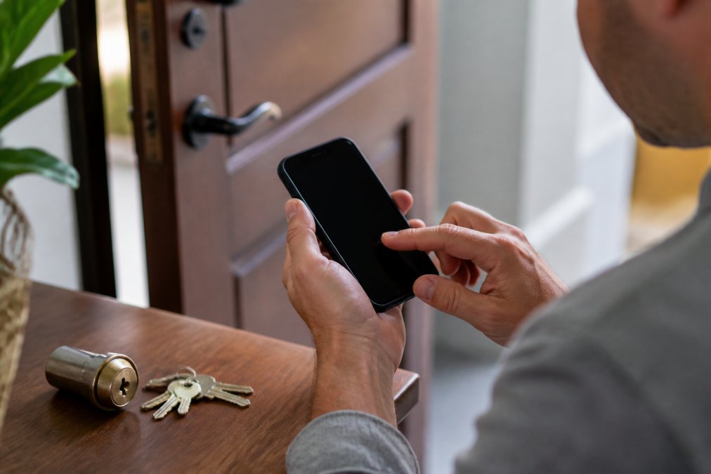 A close-up of a homeowner's hands holding a smartphone displaying a business license lookup website, with a set of keys and a lock on a table nearby in a well-lit home entryway