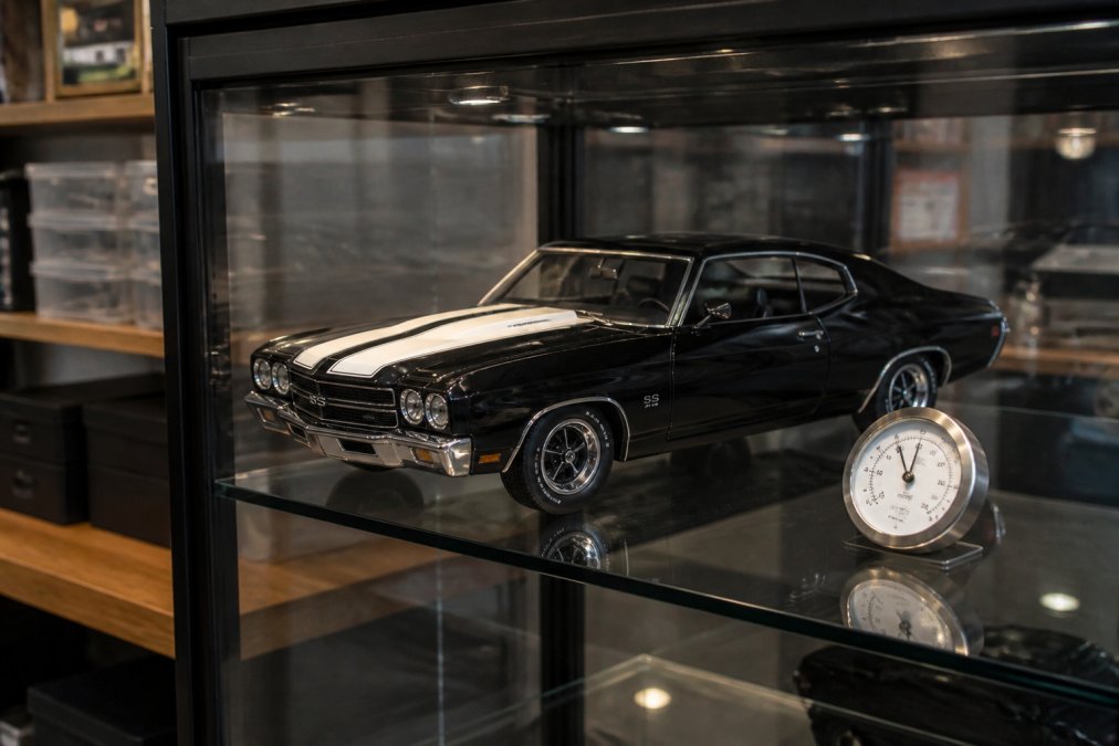 A pristine 1:18 scale diecast muscle car displayed inside a glass-front display case, with a small analog humidity gauge visible on the shelf nearby, soft ambient lighting