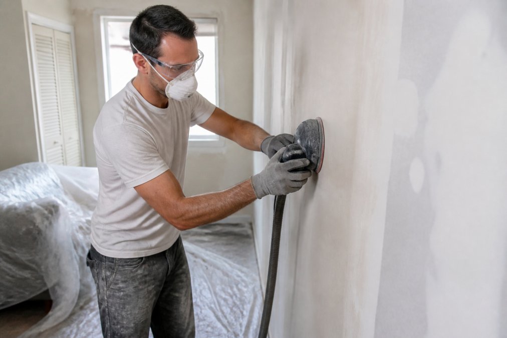 Person wearing safety gear sanding a wall covered in plastic to manage paint dust