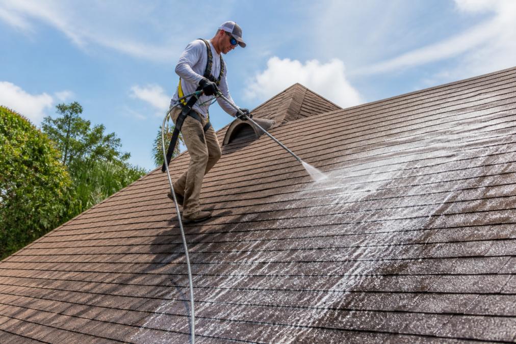 A professional technician in safety gear using a low-pressure soft wash system on a residential shingle roof, cleaning solution spray visible, no high-pressure wand, bright afternoon sunlight