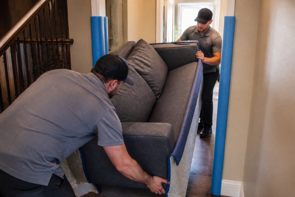 Experienced movers carefully guiding a large upholstered sofa through a narrow hallway using foam corner guards and protective padding on the doorframe while a second crew member steadies the piece