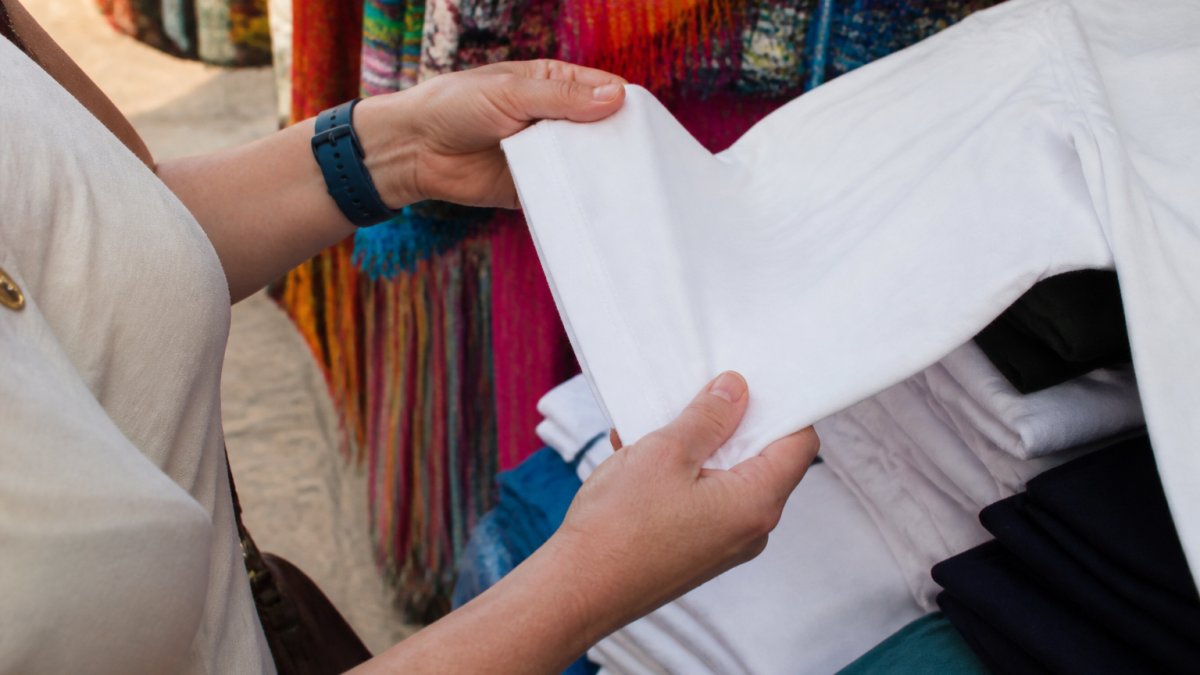 Close-up of hands examining the stitching and fabric weight of a cotton T-shirt at an outdoor market stall, with colorful textiles in the background