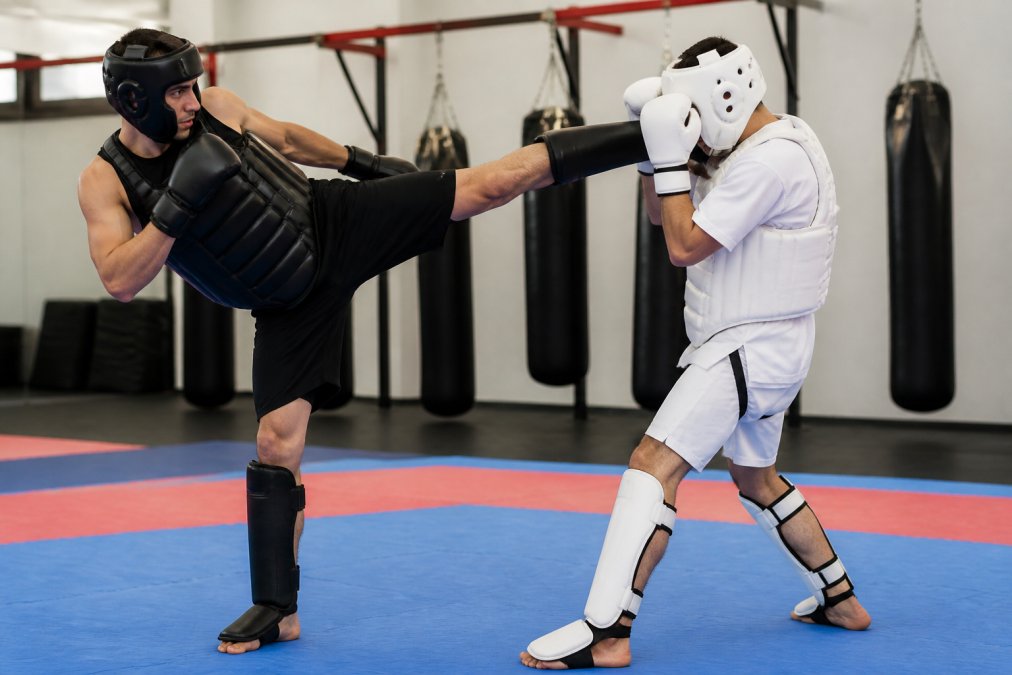 Martial artist in full protective sparring gear (padded gloves, shin guards, headgear) demonstrating proper fit and protective coverage during training drill with focused intensity