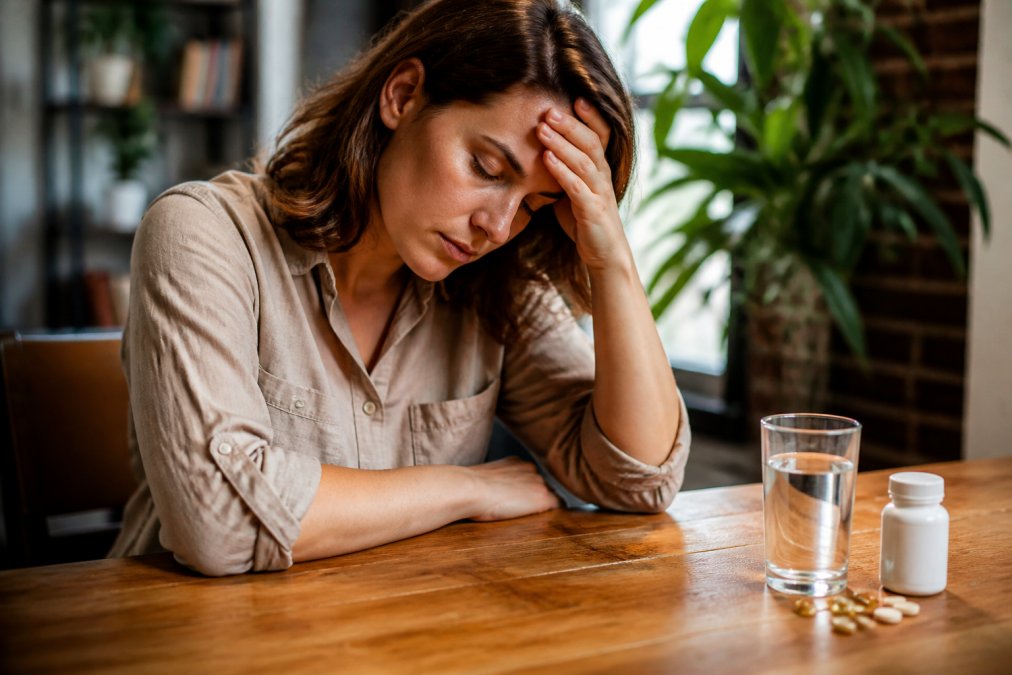 A tired adult sitting at a desk with head resting on hand, eyes closed, looking fatigued, with a glass of water and vitamin supplements nearby on a wooden desk in warm natural light