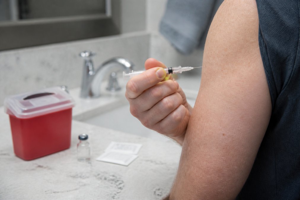 Close-up of a person's hand holding a small syringe near their upper arm, preparing for a self-injection in a clean, well-lit home bathroom, with alcohol swabs and a sharps container visible on the counter