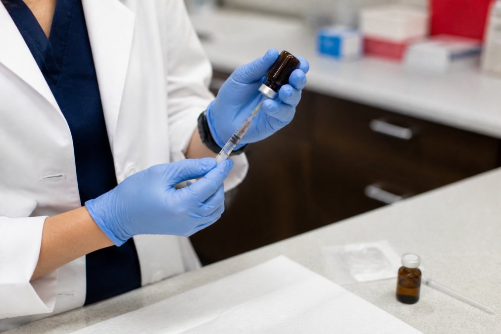 A medical professional's hands preparing a vitamin B12 injection vial and syringe on a clean clinic counter, bright overhead lighting