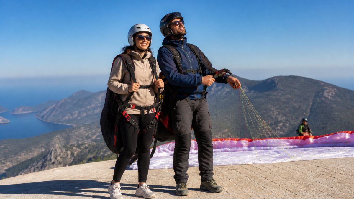 Paraşütçü pilot ve turist, uygun giyimle Babadağ'da uçuş hazırlığı sırasında, mavi gökyüzü ve dağ manzarası arka planda