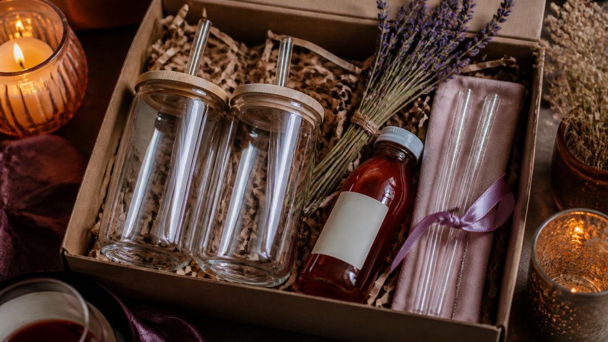 A flat-lay photo of a curated bachelorette gift box on a table, featuring two matching glass can cups, cocktail mix, reusable glass straws, and lavender sprigs, styled with warm lighting and a festive girls' night aesthetic