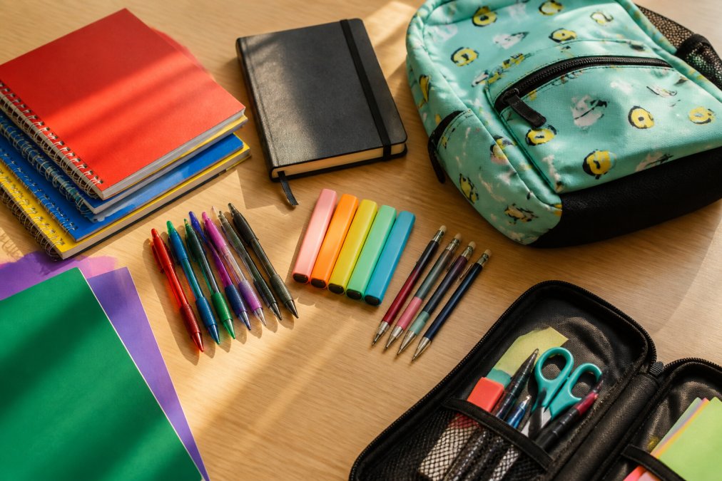 Organized collection of back to school essentials displayed on a light wooden desk: colorful notebooks, ballpoint pens in various colors, highlighters, mechanical pencils, folders, a hardcover journal, and a casual backpack with fun designs, natural morning light streaming across the items