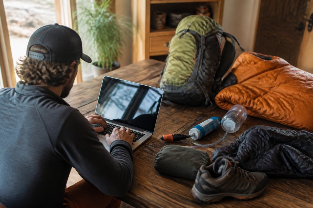 Person browsing outdoor gear selection on a laptop, surrounded by camping equipment including backpack, sleeping bag, water filtration system, and technical apparel on a wooden desk with natural window lighting