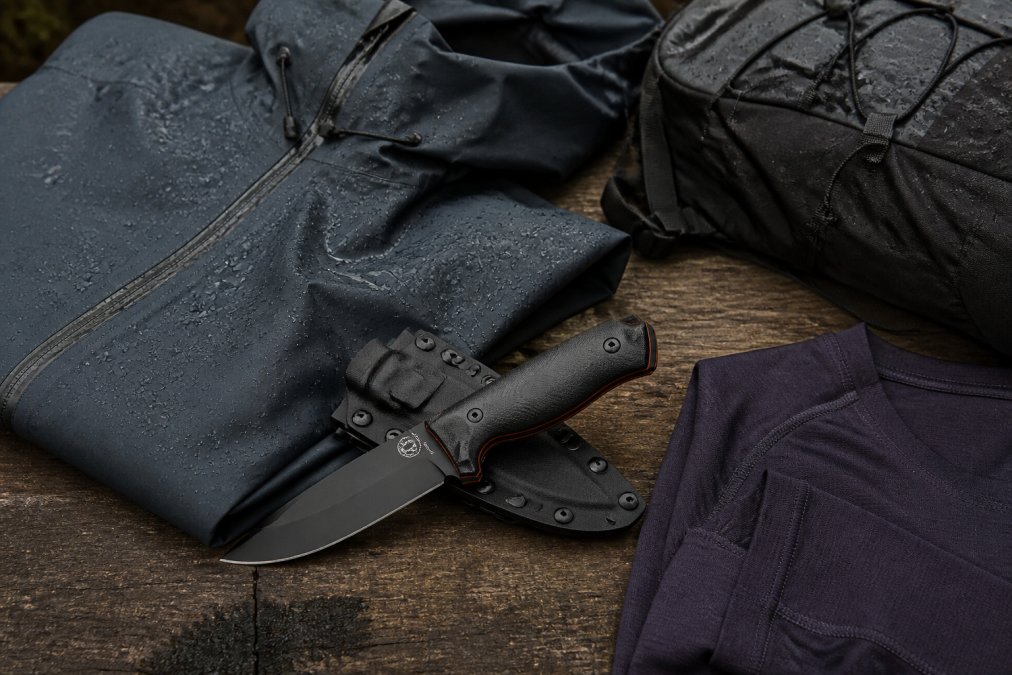 Close-up of expedition-grade gear including a weatherproof shell jacket, ultralight backpack, and technical base layers arranged on a wooden surface with visible water beads and moisture on the fabric