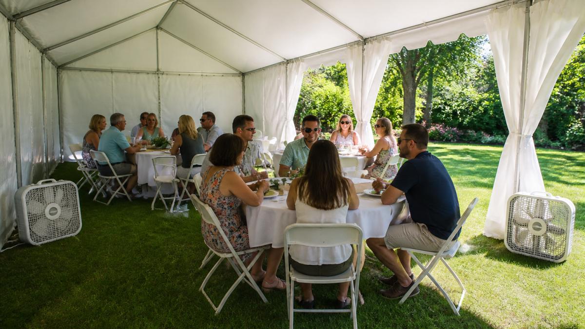 A white party tent set up in a lush backyard with sidewalls partially open on one side, two box fans placed at opposite corners, and guests in summer attire seated comfortably underneath in bright afternoon light