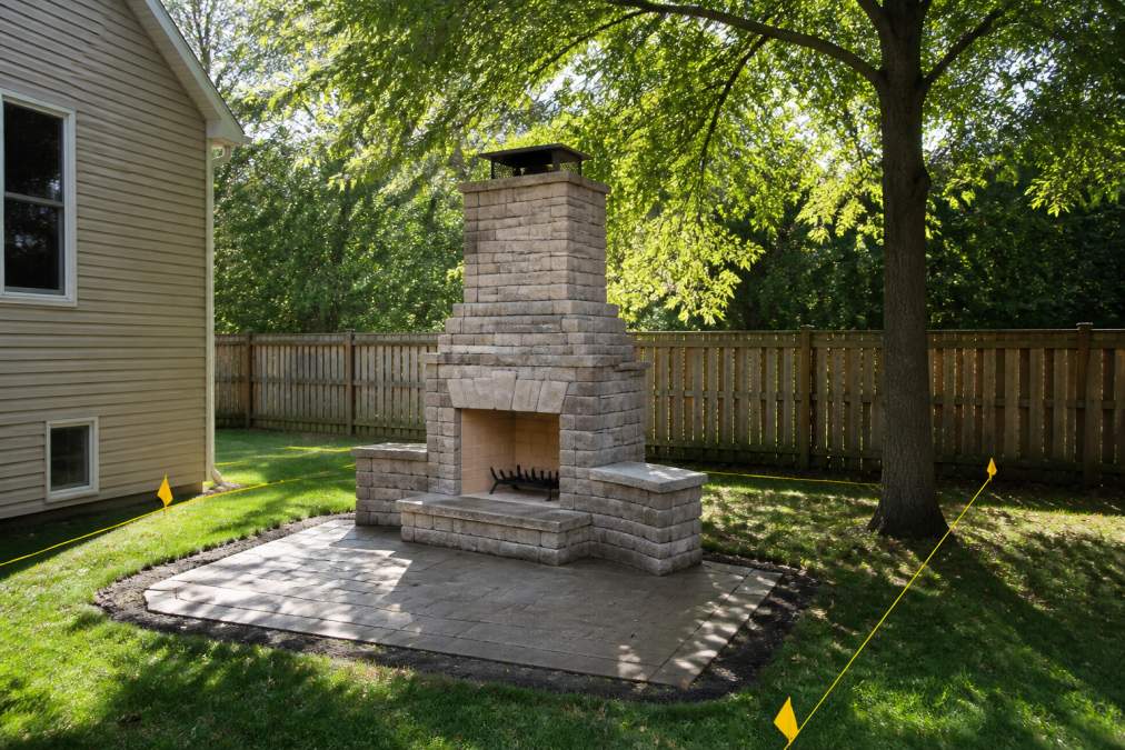 Residential backyard with permanent outdoor fireplace installation showing marked clearance zones from house wall, overhanging tree branches, and property line with safety distance markers visible in afternoon sunlight