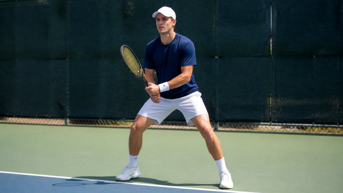 Competitive tennis player mid-split step on the court, feet shoulder-width apart, knees bent, weight balanced on the balls of the feet, demonstrating proper athletic positioning during opponent's stroke, outdoor hard court with natural daylight