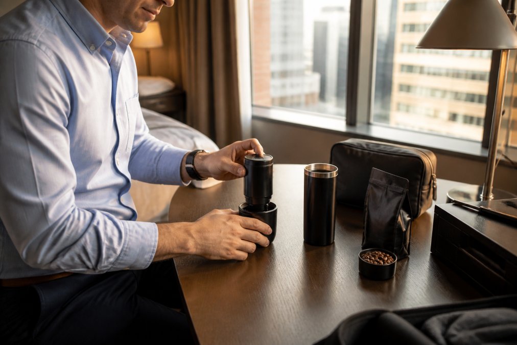 Forretningsrejsende opstiller Wacaco Nanopresso og en lille pose kaffebønner på et hotelskrivebord, morgenlys strømmer ind gennem gardiner, sort rejsetaske åben ved siden af
