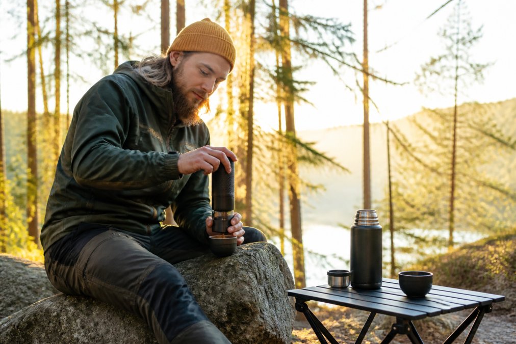 Person i outdoor-tøj brygger kaffe med en kompakt bærbar kaffemaskine på en sten i den smukke morgenmadsnatur.