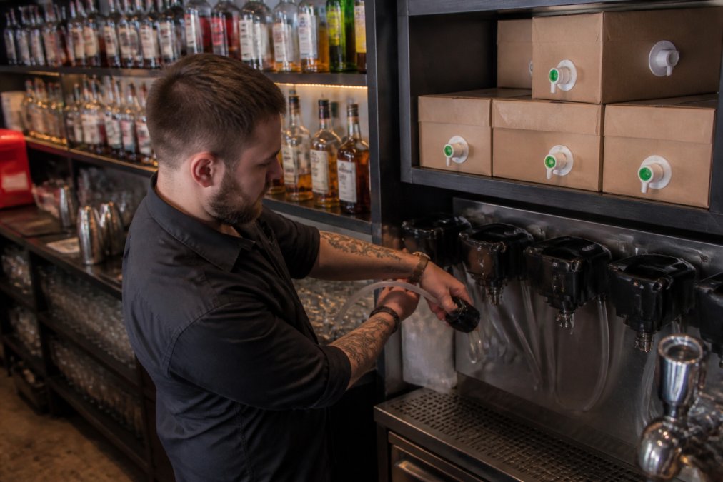 A bartender in a busy pub connects a fresh bag in box syrup container to the post-mix dispenser behind the bar, with a neat stack of spare boxes on the shelf behind