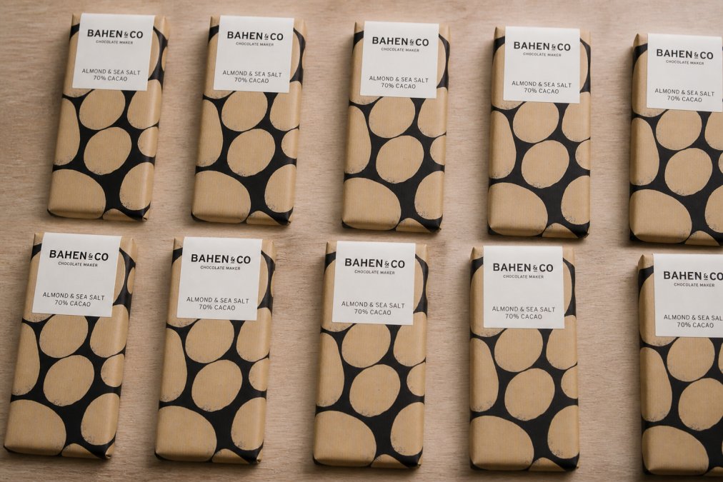 Bahen and Co chocolate bars arranged in rows on a light wooden surface, showing the variety of flavour labels and rich cocoa tones, with soft natural light highlighting the texture and artisanal craftsmanship of each bar