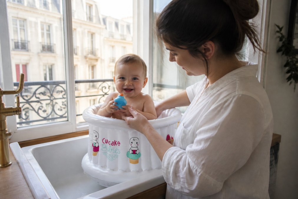 Parent tenant un bébé dans une petite baignoire gonflable Cupcake Babies posée dans un évier de cuisine moderne, lumière naturelle du jour, appartement parisien avec salle de bain compacte