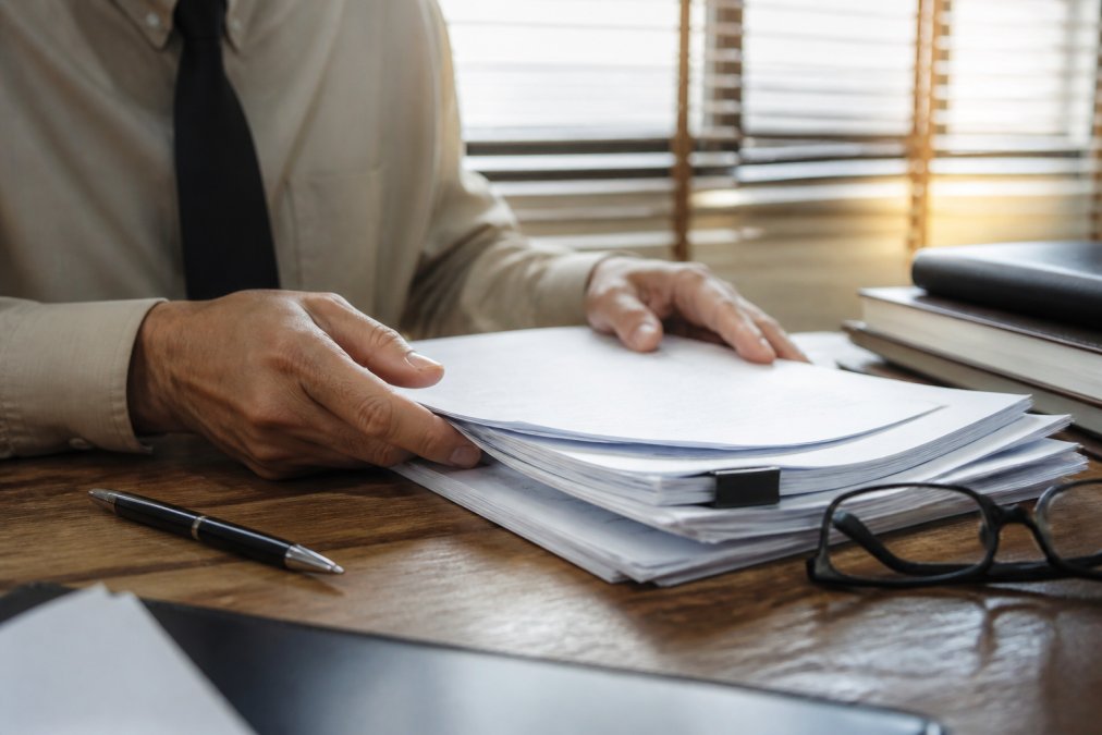 Close-up of hands reviewing a stack of legal documents on a wooden desk, pen and reading glasses nearby, warm office lighting through window blinds