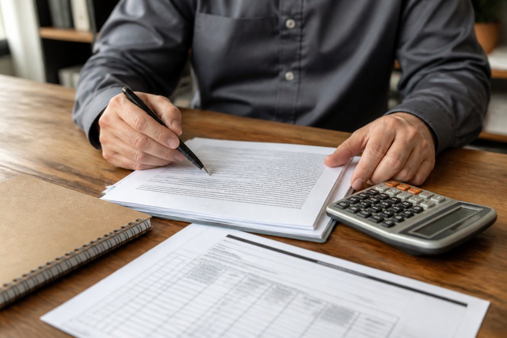 A close-up of a person reviewing a bail bond contract with a pen in hand, sitting at a wooden desk in a professional office setting, with a calculator and paperwork visible
