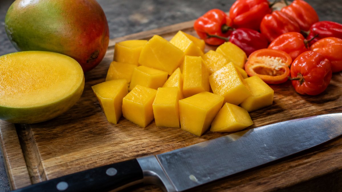 Close-up of fresh mango and habanero peppers on a cutting board with a chef's knife, showing prepared fruit chunks ready for roasting