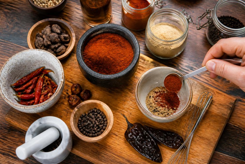 Close-up overhead shot of various spice bowls and jars (smoked paprika, chipotle powder, black cardamom, whole peppers) arranged on a wooden cutting board with a hand measuring spices into a small bowl