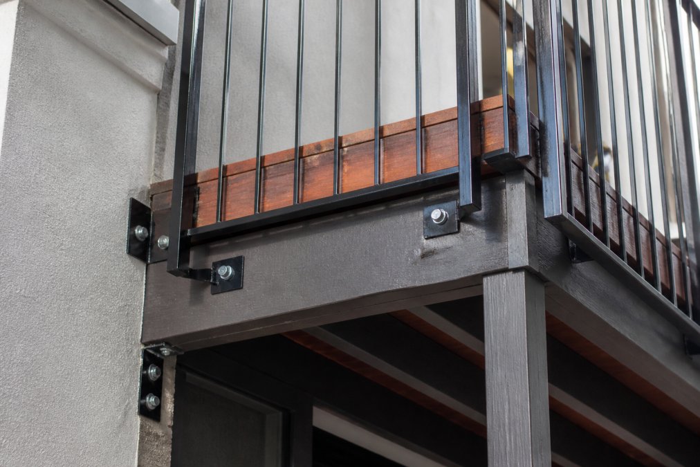Close-up of a residential balcony with wooden decking, metal railings, and structural support beams visible in natural daylight, showing the connection points where the balcony attaches to the building
