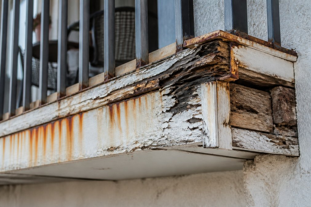 Close-up of severely deteriorated balcony railing with visible rust stains, peeling paint, and water-damaged wood framing showing deep cracks and wood rot discoloration