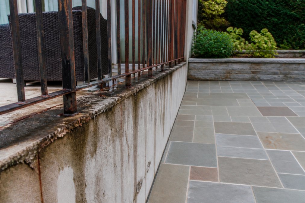 Close-up of a weathered balcony railing with visible corrosion on metal balusters and water staining on the building wall below, positioned next to a well-maintained stone patio surface with clean grout lines and no visible deterioration