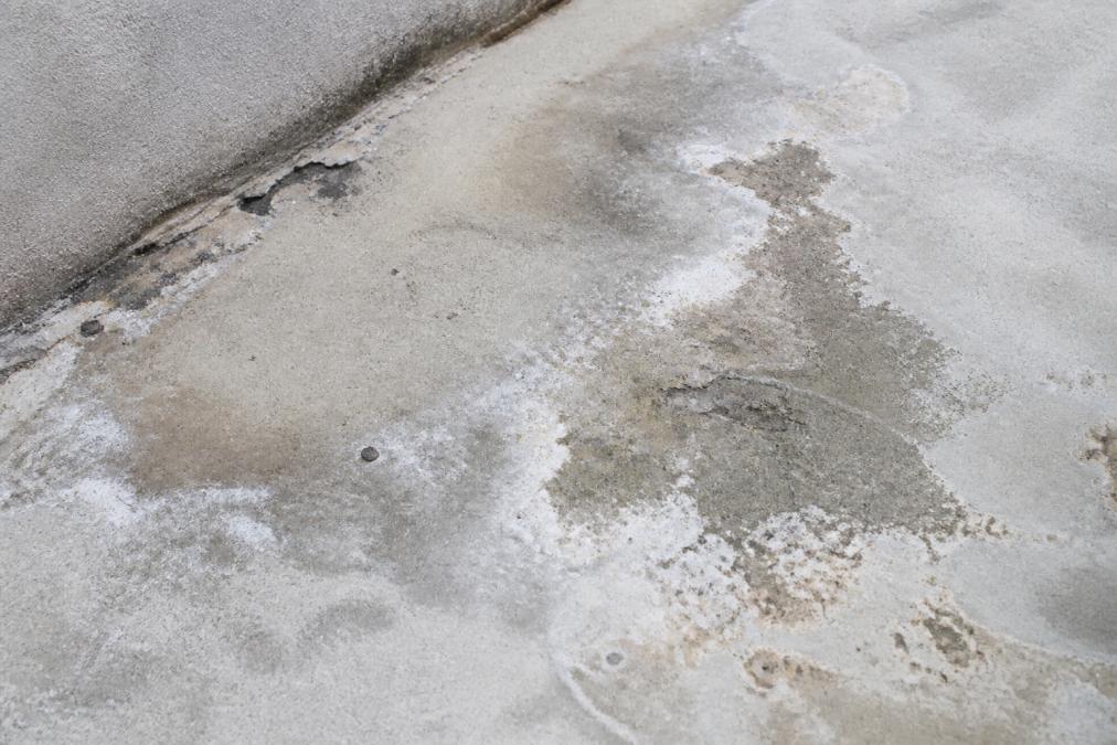 Close-up of concrete balcony surface showing visible water staining, white efflorescence deposits, and early spalling with moisture marks and discoloration patterns