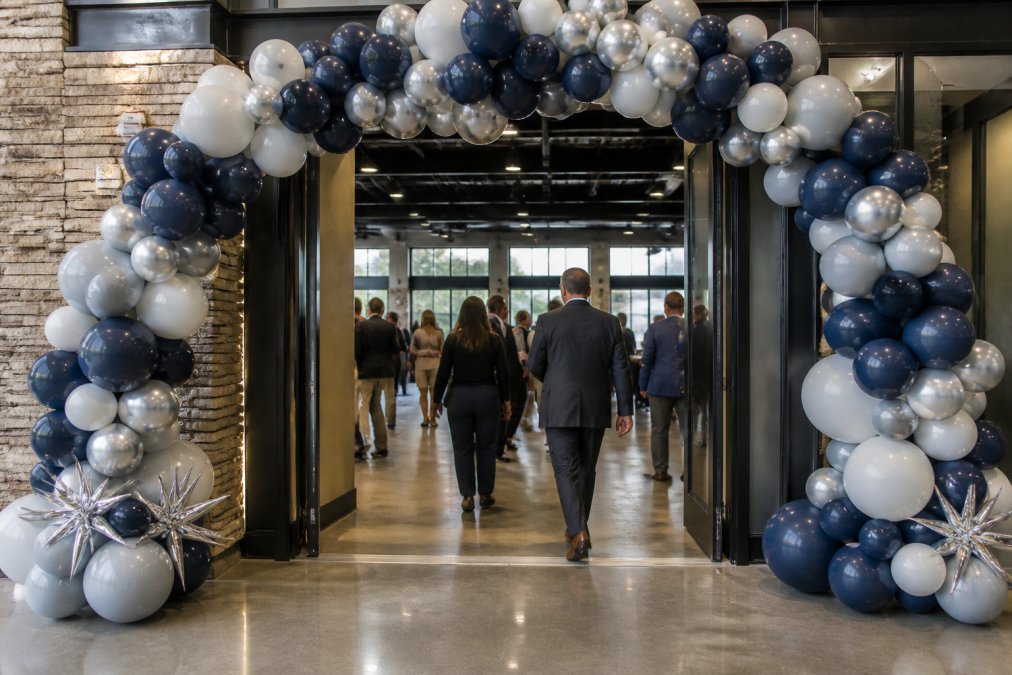 A large custom balloon arch in corporate brand colors framing the entrance to a professional event venue, with guests walking through in business attire