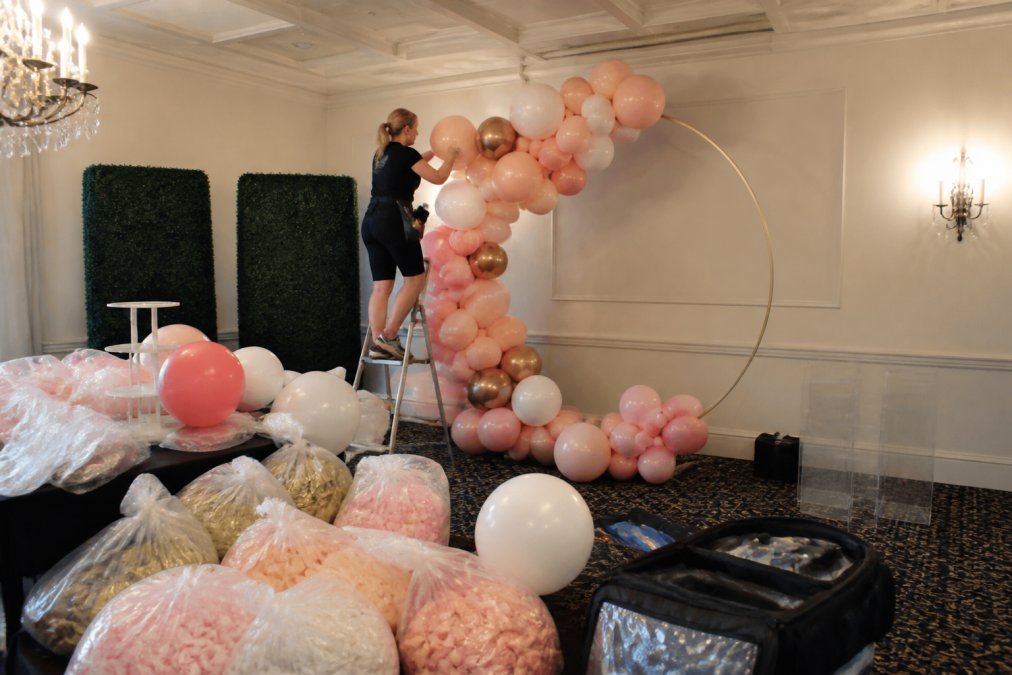 Professional balloon decorator installing a large organic balloon arch at an event venue, carefully arranging balloons in various colors and sizes on a metal frame structure, with additional balloon materials and tools visible in the foreground