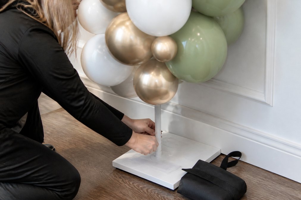 Close-up of professional hands carefully attaching balloon weights to a balloon arch base, using adhesive strips and proper fastening techniques on an interior wall