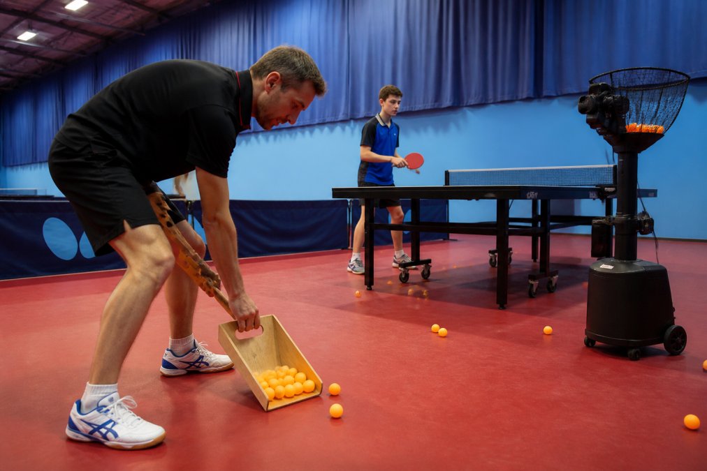 A table tennis coach using a handheld wooden scoop to gather orange ping pong balls from a gym floor, with a multi-ball robot and a junior player practicing at a table in the background under bright overhead lighting