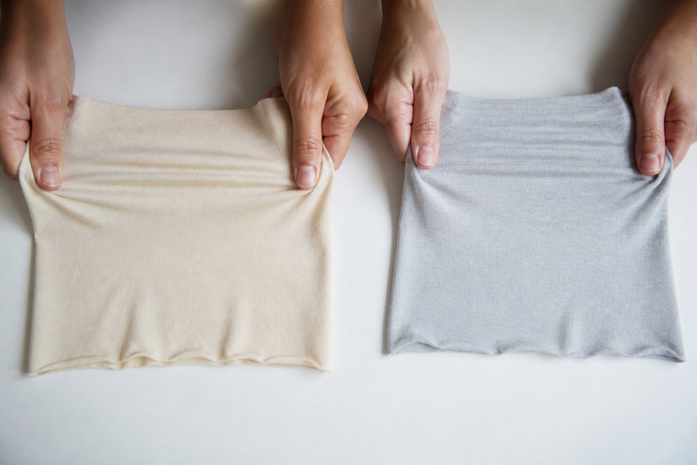 Person's hands gently stretching two fabric samples side-by-side on a white surface, demonstrating elasticity and fiber recovery, with one fabric bouncing back more quickly than the other, natural daylight illuminating the textures