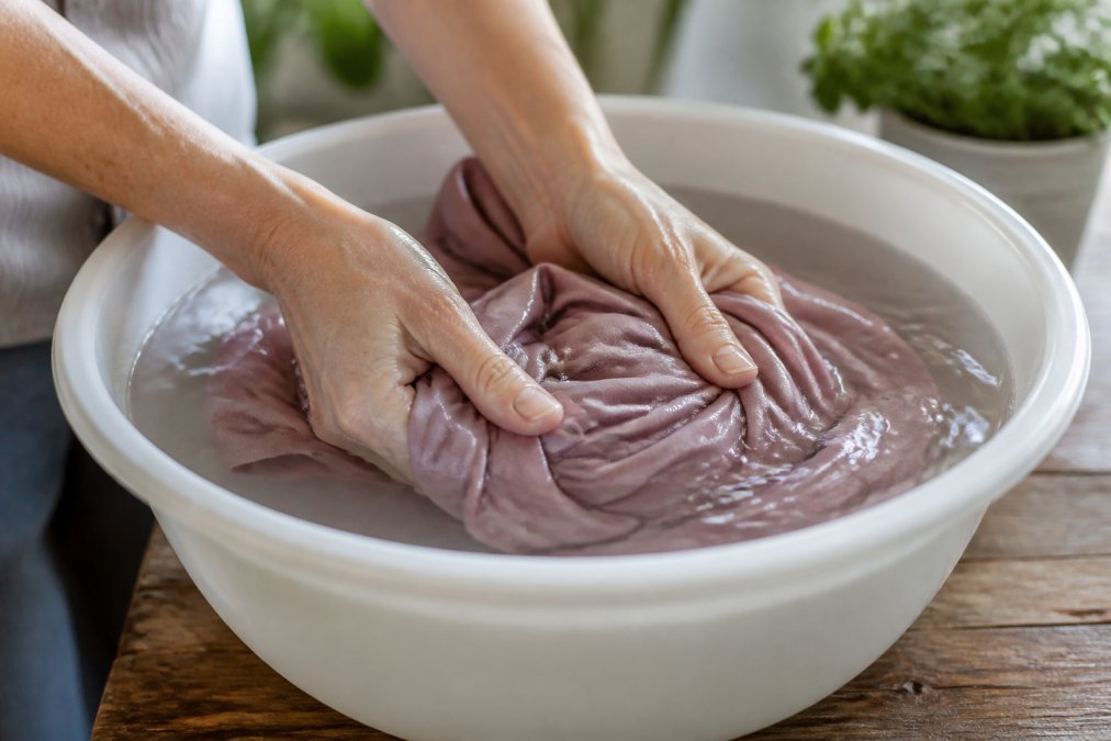 Hands gently washing a modal dress in cool water, showing proper hand-washing technique with the fabric submerged in a white basin, demonstrating careful care