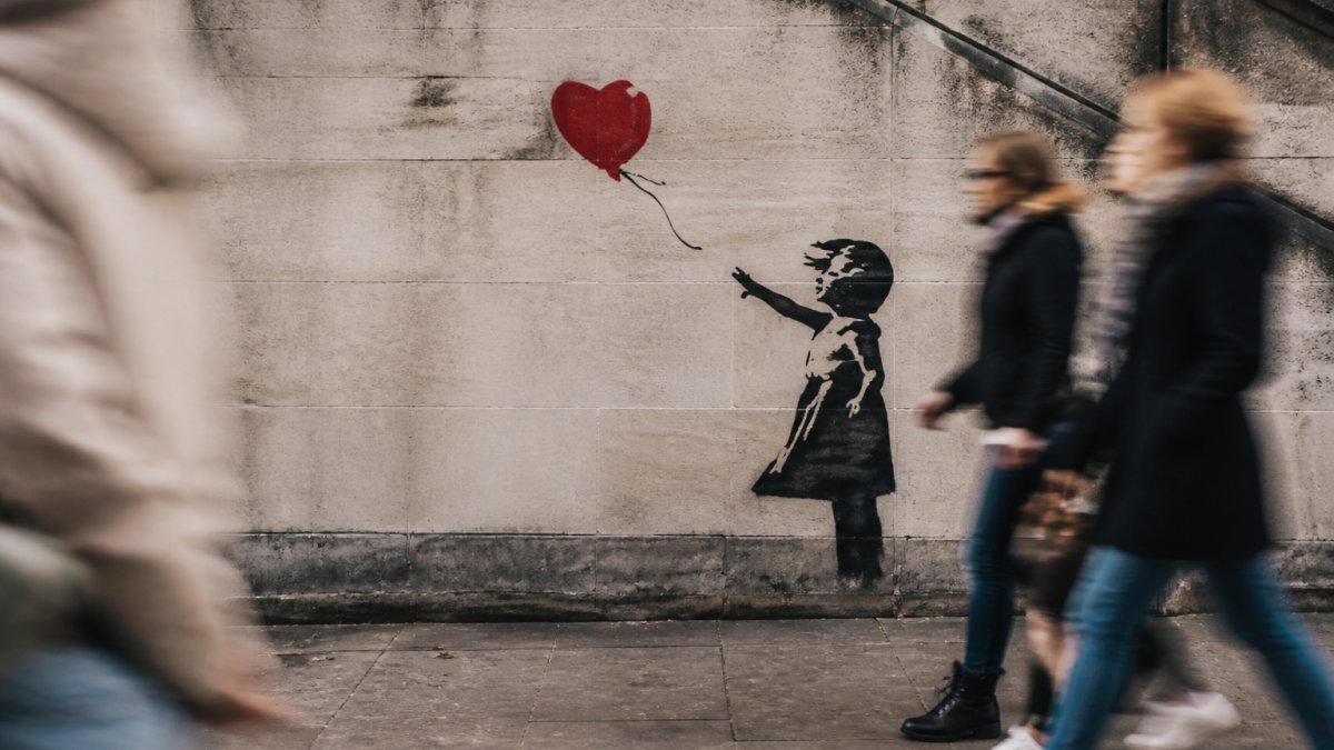 A close-up photograph of a weathered concrete wall featuring a stencil of a girl reaching for a red heart-shaped balloon, with passersby blurred in the background on a city street