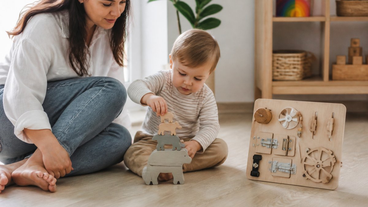 A parent and toddler sitting on a light wood floor, the toddler stacking a solid wood animal puzzle while a wooden busy board sits nearby, soft natural window light, calm home playroom setting