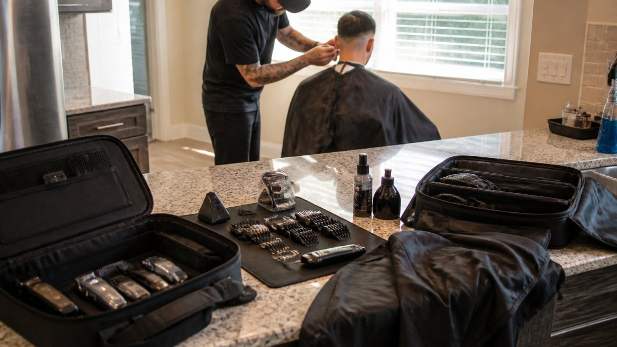 Professional barber setting up portable equipment on a client's kitchen counter with organized clipper cases, water-resistant cape, and tools laid out systematically, natural sunlight streaming through window