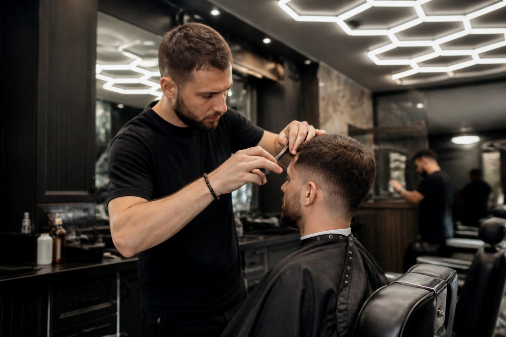 Un barbier concentré qui coupe les cheveux aux ciseaux dans un salon au décor moderne, éclairage hexagonal au plafond, client assis dans un fauteuil de barbier en cuir