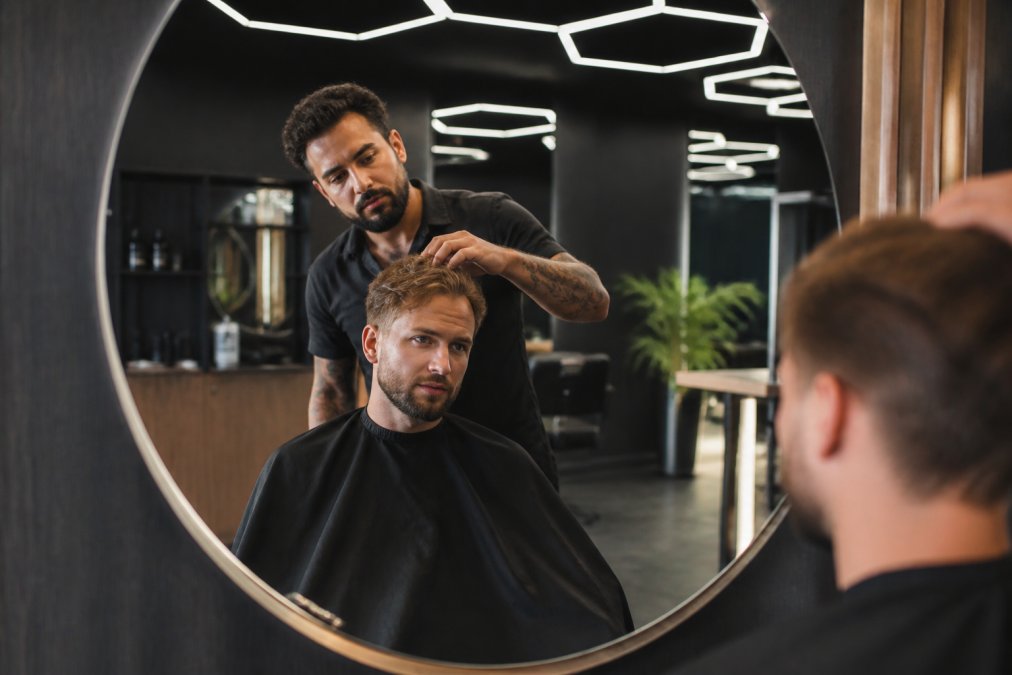 Un barbier et son client en consultation devant un miroir de salon, le barbier touchant délicatement les cheveux du client pour évaluer la texture, dans un espace moderne avec un éclairage hexagonal distinctif