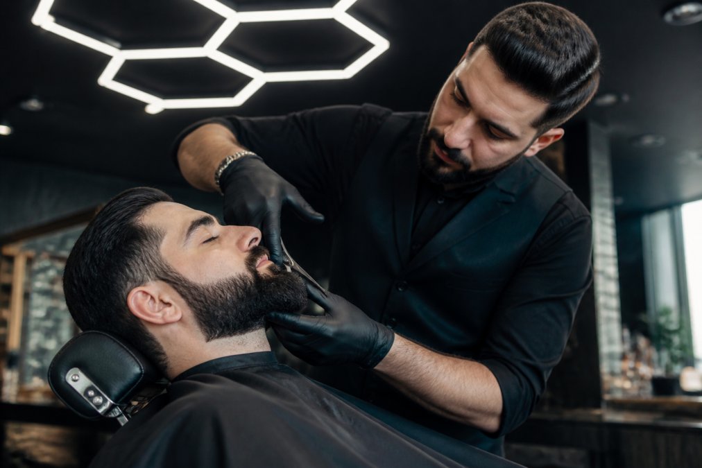 Client relaxing in a luxurious leather barber chair in a modern salon with distinctive hexagonal lighting overhead, while a skilled barber in professional attire carefully works on sculpting his beard with precision scissors