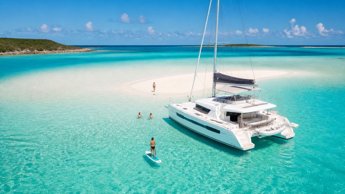 Aerial view of a luxury catamaran anchored in a shallow turquoise Bahamas sandbar with white sand and a few people swimming and paddleboarding nearby