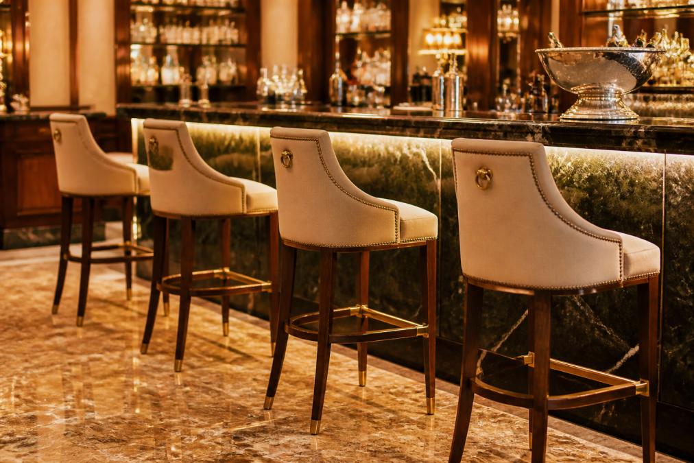 Elegante Barhocker aus Massivholz mit aufwendiger Polsterung an einer Marmor-Theke in einem luxuriösen Hotel-Barbereich, warmes Licht, edle Innenausstattung