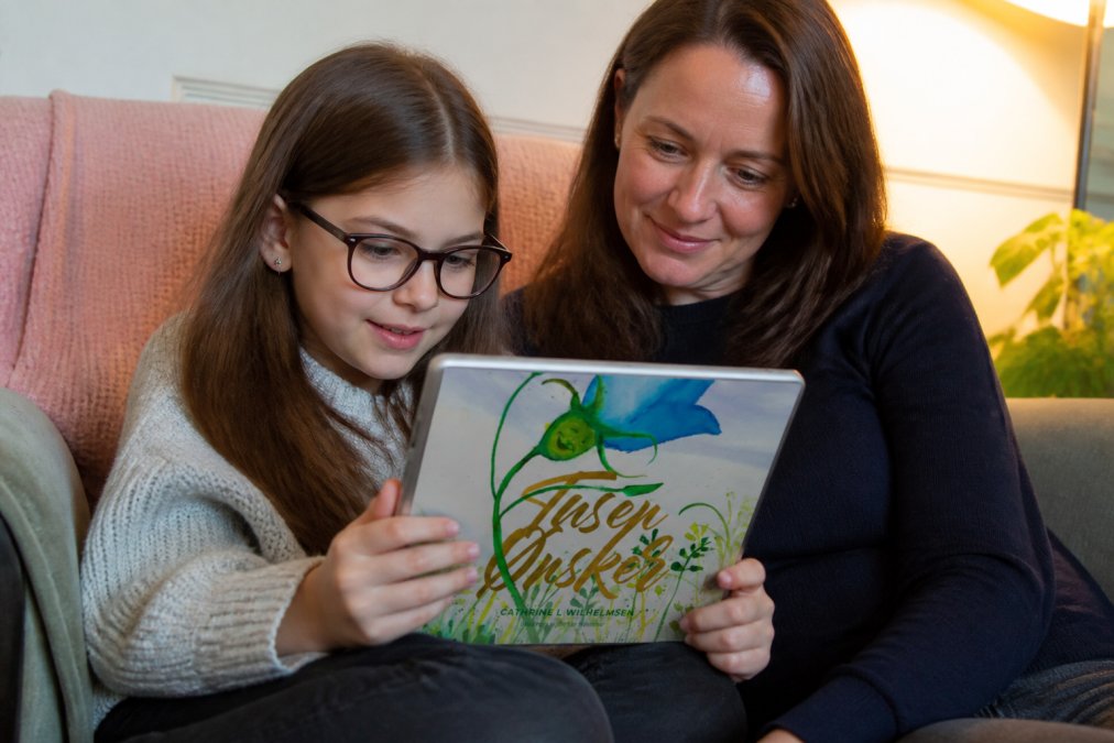 Et barn sitter komfortabelt og leser en illustrert bok i et koselig hjørne med god belysning, mens en voksen sitter ved siden av og lytter oppmerksomt